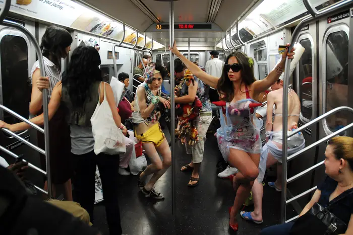 Inside a semi-crowded L train long before COVID-19, people in weird costumes dance through the subway car.